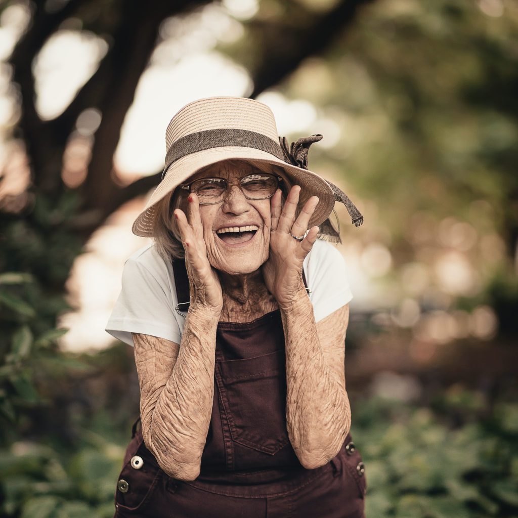 An old woman with a bunch of wrinkles wearing a straw hat. Her hands are on the sides of her face and she looks like she is yelling at some. Smiling real big | who does god say I am | Positively Jane