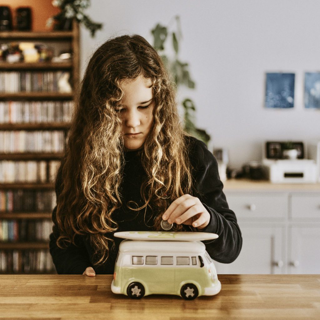 Small girl with long curly hair putting money in a piggy bank. The bank is shaped like a VW bus with a surf board on top | 10 reasons you need an emergency fund | Positively Jane