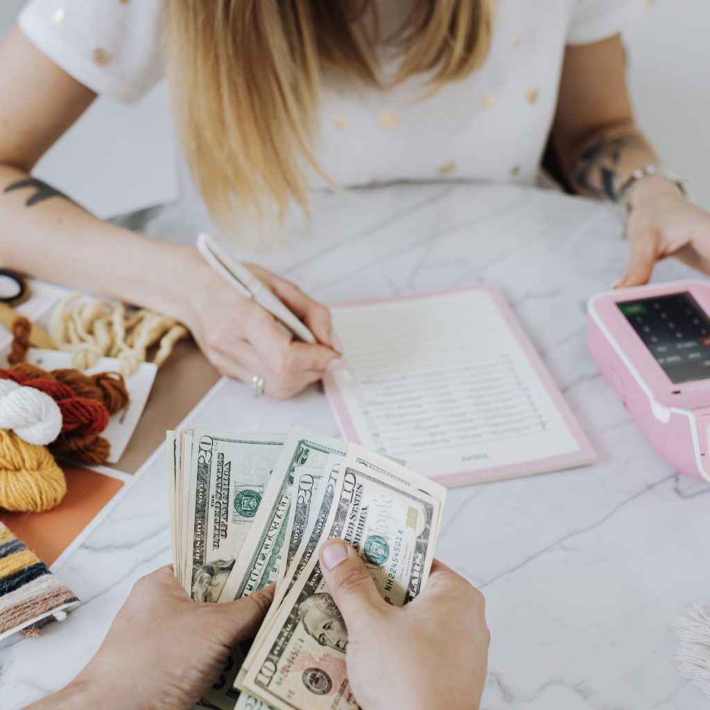 Woman with a calculator and a list. Another person with money in their hands |  how to establish budget categories |Positively Jane