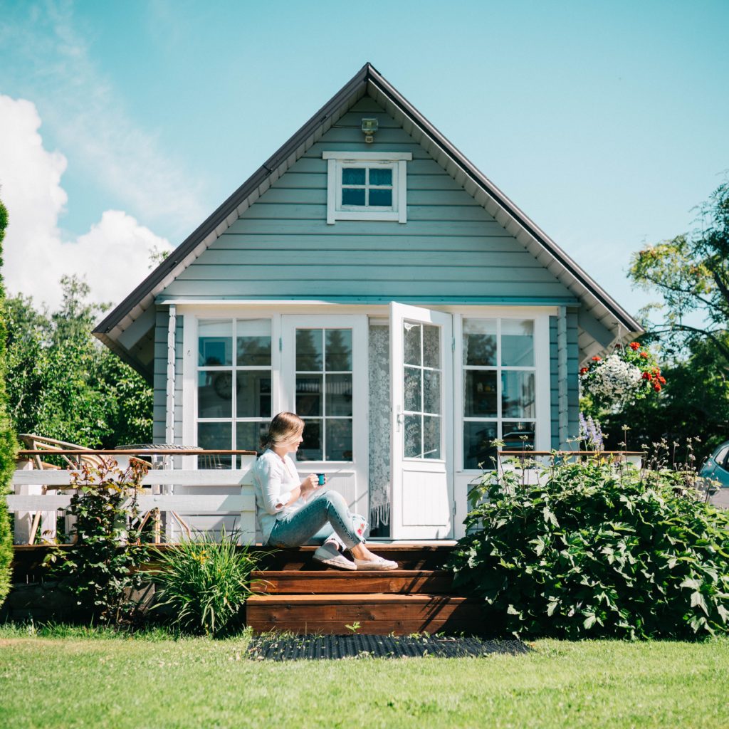 small blue house with white windows and doors. A deck on the side and a girl sitting on the deck | saving to buy a house | Positively Jane  