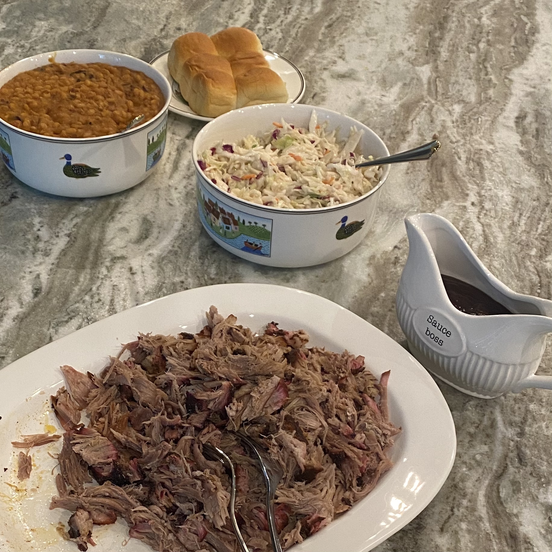 pulled pork on a plate, a bowl of beans, a bowl of coleslaw, a gravy boat with barbecue sauce and buns