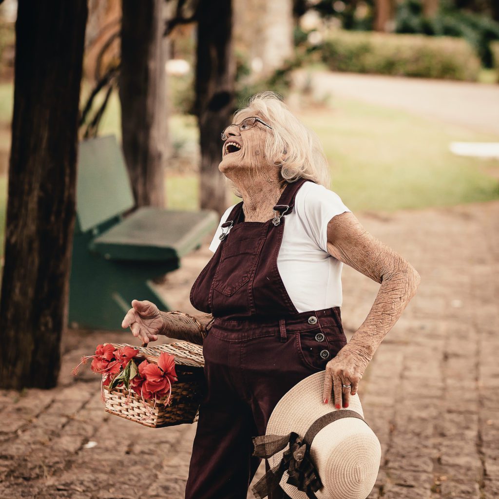 old woman with many wrinkles. She has her head thrown back and is laughing out loud. She is holding a straw hat in one hand and flowers in the other | finding joy in all things | Positively Jane