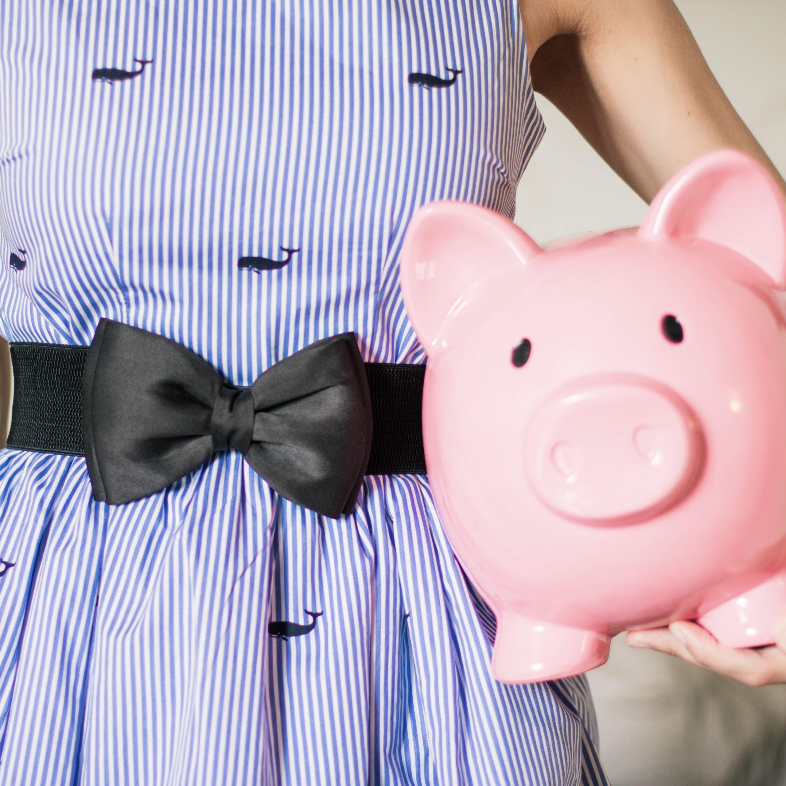 girl in purple and white striped dress with a black satin bow in front holding a large pink piggy bank | how to teach kids about money | Positively Jane