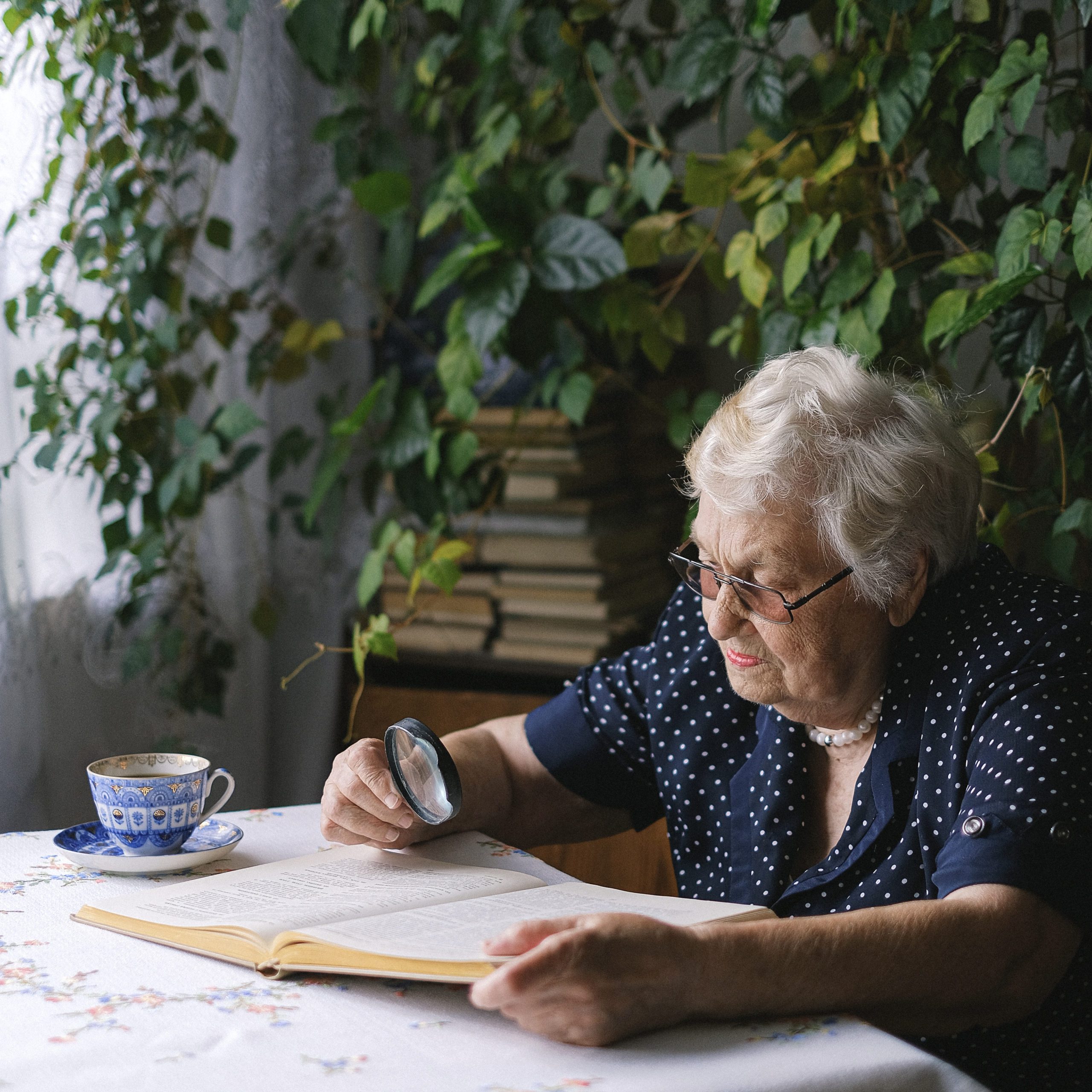 older woman reading a book with a magnifying glass. She is sitting at a table. There is a long viney plant growing behind her | saving for retirement | Positively Jane